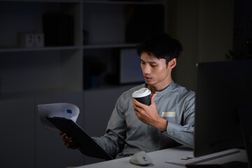 Serious male employee focused on reading report with hot coffee in hand at dark office