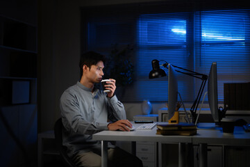 Asian businessman working late night at desk, drinking coffee to stay awake in front of computer