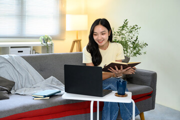 Casual woman reading book and taking notes while working on laptop at home
