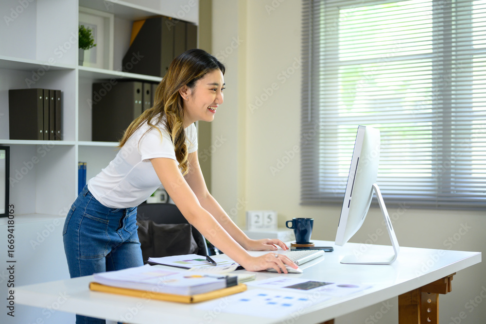 Poster happy young woman analyzing data and reports while working on desktop computer at desk