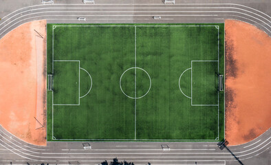 Aerial top view of an empty football soccer field with green artificial grass and white marking lines, surrounded by a running track. Symmetrical sports background, perfect for illustrating teamwork, 