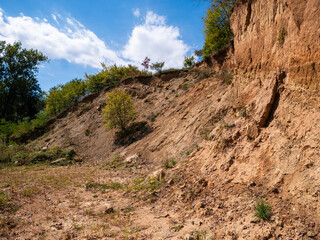 Exposed sandy and clay hillside with signs of erosion, vegetation on top, and clear sky background.
