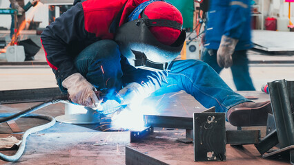 A skilled worker is focused on welding metal pieces together in a fabrication shop. Bright sparks...