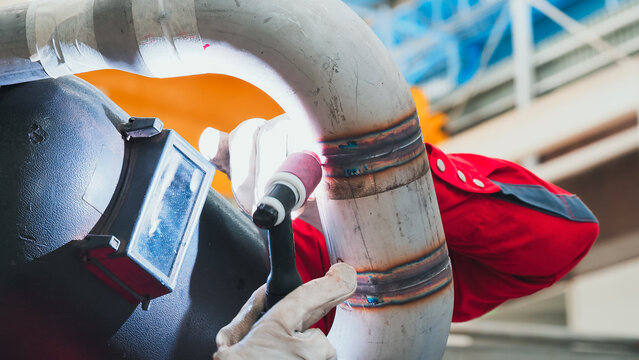 A technician in a bright red uniform works diligently on welding a metal pipe in an industrial setting. Bright lights illuminate the workspace, showcasing attention to detail and craftsmanship