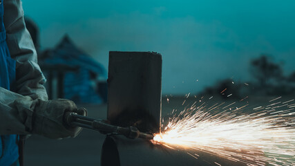 A welder operates a gas torch in an industrial workshop, producing bright sparks while working on metal fabrication. The setting is dimly lit, emphasizing the intense activity
