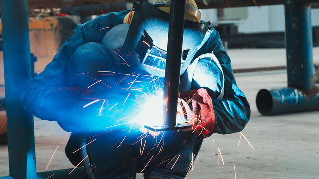 In an industrial workshop, a skilled welder crouches down, focused on joining metal pieces. Bright blue sparks fly as the welding torch fuses materials together. Safety gear ensures protection