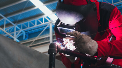 A welder is engaged in a task at an industrial location, concentrating on joining metal pieces with intense focus. Protective gear ensures safety while performing the intricate work