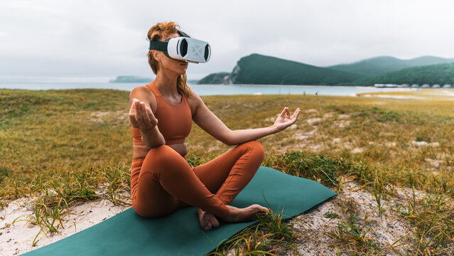 Woman in sportswear sits on yoga mat with VR headset, meditating outdoors near sea. Concept of mindfulness, digital wellness, balance between technology and inner peace. - Powered by Adobe