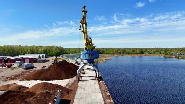 From a high drone, a bulk-handling port crane straddles a concrete quay beside calm blue water, with brown stockpiles, service buildings, and forest fringe under crisp, breezy daylight.