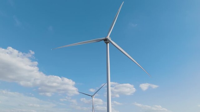 Close-up drone shot of a tall wind turbine set against a bright blue sky with clouds.