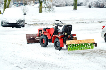 Red tractor snow removal machine with brush attachment clearing road during winter season on parking area, outdoor maintenance equipment, snow clearing vehicle with rotary brush, compact red tractor