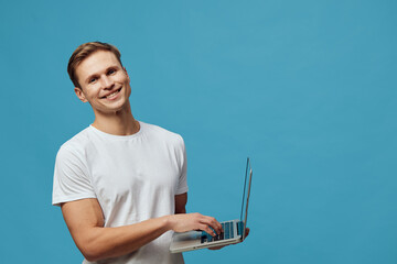 Smiling young man holding a laptop computer with one hand, standing against a solid blue background...