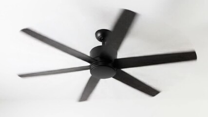 A black ceiling fan with six blades is spinning creating a blurred effect of motion against a white ceiling