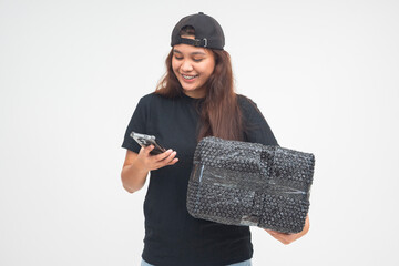 Cheerful young boyish woman checking her phone while holding a black parcel for delivery. Isolated...