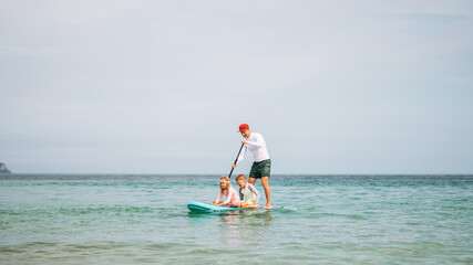 Dad and kids paddle together on SUP board during beach holiday. Summer joy, ocean adventure, and active lifestyle captured in cheerful family moment full of energy and smiles.