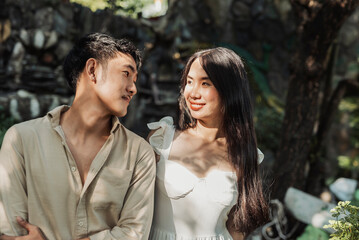 A happy young Southeast Asian couple standing closely under tree shade while smiling warmly and gazing into each other's eyes with affection.