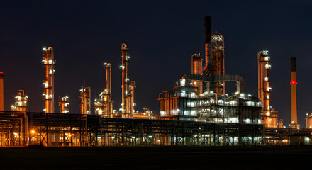 Fototapeta premium Illuminated industrial complex at night with multiple towers and pipelines under a dark blue sky, creating a stark contrast between the bright facility and the surrounding darkness.