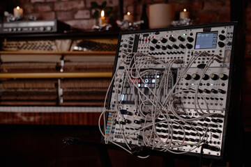 Modular synthesizer with numerous patch cables connected, standing in front of upright piano with exposed strings and brick wall background, electronic music production equipment