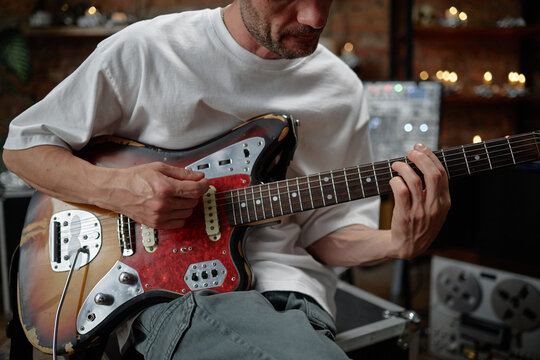 Caucasian man playing electric guitar, focusing on hand positioning and finger technique, sitting indoors with visible music equipment in background