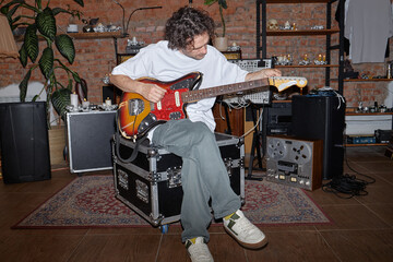 Caucasian man sitting on equipment case tuning electric guitar in music studio surrounded by audio gear and vintage electronics, focusing on instrument adjustment