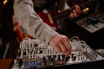 Caucasian man adjusting modular synthesizer while playing electric guitar, focusing on hand turning knob with visible instrument cables and blurred background