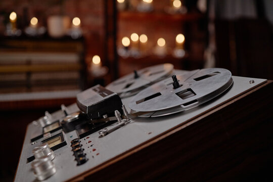 Closeup showing vintage reel to reel tape recorder with large metal reels in focus, blurred background with glowing candles creating soft bokeh