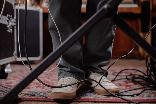 Man standing on patterned rug surrounded by audio cables and microphone stand in music studio, only lower legs and feet visible, wearing casual shoes