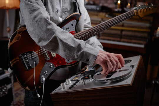 Caucasian man adjusting reel to reel tape recorder while playing electric guitar, focusing on hands and upper body, musical equipment visible in background