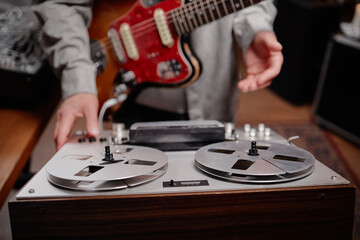 Caucasian man operating vintage reel to reel tape recorder while playing electric guitar, focusing on adjusting controls and interacting with audio equipment in music studio