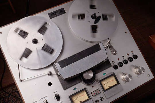 Vintage reel to reel tape recorder operating on wooden surface, large metal reels spinning while analog VU meters displaying audio levels, classic audio equipment in use