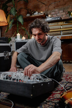Caucasian man sitting on patterned rug adjusting modular synthesizer equipment with focused expression, surrounded by musical instruments and vintage decor elements