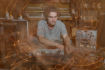 Caucasian man sitting on floor operating modular synthesizer surrounded by vintage audio equipment and open upright piano with light trails creating dynamic visual effect