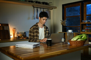 Fototapeta premium Focused man working late at night on laptop in kitchen with coffee