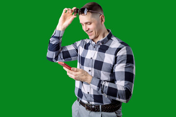 A young man in a long-sleeved plaid shirt, a braided belt and sunglasses against a green screen. He holds a phone and smiles while looking at the screen.