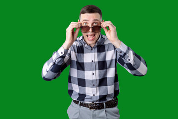 Young man in checkered shirt and braided belt holding sunglasses with surprised expression on face against green screen background.