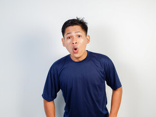 A young Asian man wearing a blue shirt expresses shock or surprise, standing against a plain...