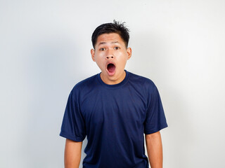 A young Asian man wearing a blue shirt expresses shock or surprise, standing against a plain...