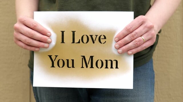 A Woman&rsquo;s Hands Holding Up a Gold Stencil Sign That Says I Love You Mom in front of a Tan Backdrop 4K Video
