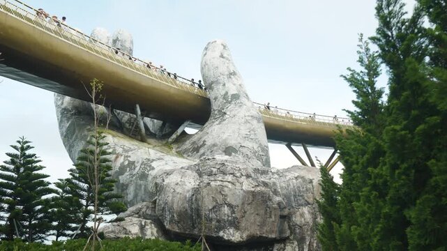 Golden bridge spanning rocky terrain, supported by giant stone hands, with visitors walking and admiring panoramic landscape at ba na hills mountainous resort in central vietnam