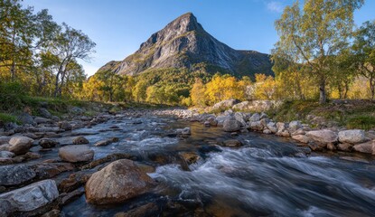 Mountain peak over flowing stream. Autumn foliage colors the trees near the water. Bright, sunny sky above