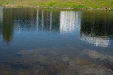 reflective lake mirroring tall buildings against a grassy bank under clear skies, highlighting contrast between nature and urban structures with vivid colors and tranquil atmosphere, translucent