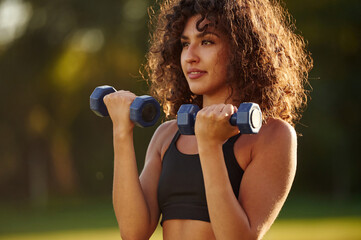Lifting the dumbbells. Young fitness woman is on the field outdoors