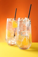 Refreshing soda water with orange slices and ice cubes in glasses on yellow table against orange background