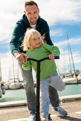 Man in hoodie teaching blond child steering scooter beside row of white boats