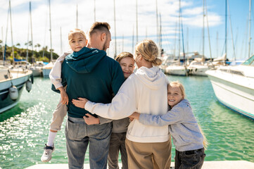 Young adult man woman and their children standing backwards to camera near yachts