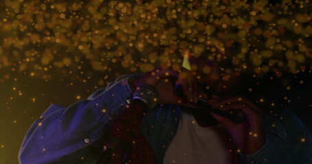 Playing musician in denim jacket blowing harmonica at microphone stand on dark stage, golden bokeh