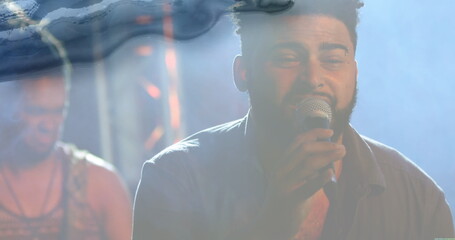 Singing bearded lead singer holding microphone on dimly lit stage, with foggy haze and metal truss