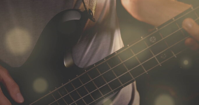 Strumming musician with guitar strap playing black bass guitar on dim stage, with lens flares