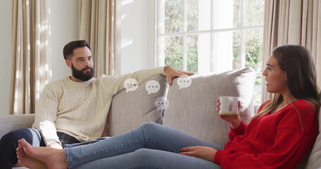 Talking couple reclining on light gray sectional sofa in living room, holding ceramic mug by window