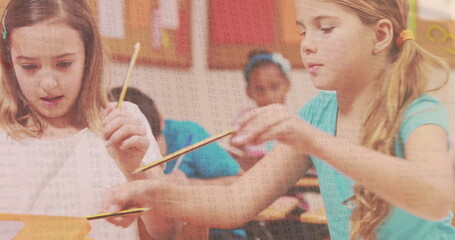 Leaning two girls writing on worksheets at wooden desks in classroom, with pencils and posters